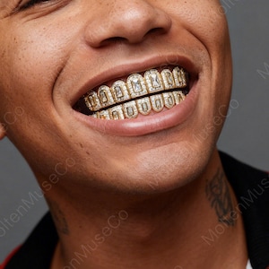 Puede incluir: Primer plano de una persona sonriendo, mostrando grillz dorados. Los grillz presentan un diseño rectangular con piedras claras y brillantes. Los dientes están completamente cubiertos con el diseño de oro y piedras.