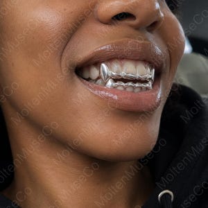 Könnte beinhalten: Ein Set aus silberfarbenen Metall-Zahngrillz, die die oberen und unteren Vorderzähne bedecken. Die Grillz haben eine glatte, polierte Oberfläche mit spitz zulaufenden Eckzahnabschnitten und sind so geformt, dass sie über die Zähne passen.