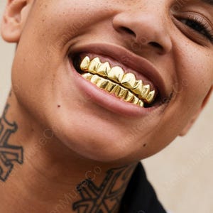 Può includere: Primo piano di un sorriso che mostra grillz dorati. I grillz sono molto lucidi, catturando la luce e creando un effetto visivo sorprendente. I denti sono perfettamente allineati, migliorando l'estetica complessiva.