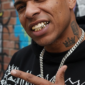 Könnte beinhalten: Nahaufnahme einer Person mit goldenen Zahnspangen, einer silbernen Kette und einem schwarzen Kapuzenpullover. Ein Mikrofon- und Kronen-Tattoo ist am Hals zu sehen. Der Hintergrund ist eine Backsteinmauer mit Graffiti.