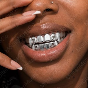 Könnte beinhalten: Ein Set aus silberfarbenen Zahngrillz, die die oberen und unteren Zähne bedecken. Die Grillz haben eine glatte, polierte Metalloberfläche mit kleinen, markanten herzförmigen Aussparungen an den Eckzähnen als dekoratives Accessoire.