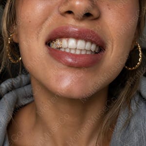 May include: A gold tooth grill cap featuring a small, clear, round-cut gemstone set in the center. The decorative piece is fitted onto a single upper tooth, showcasing a polished metallic finish and a detailed, textured border surrounding the stone.