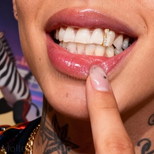 May include: Close-up of a person's mouth with a gold tooth grill. The grill is rectangular with a small pendant and embedded with small, clear stones. The person's lips are glossy and pink, and the teeth are white.
