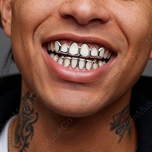Può includere: Primo piano di una persona sorridente, che mostra grillz argentati sui denti superiori e inferiori. I grillz hanno una finitura metallica lucida, in contrasto con i denti naturali. È visibile l'incarnato della persona, insieme a un tatuaggio sul collo.