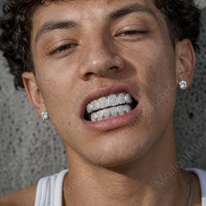 Può includere: Primo piano di una persona che indossa grillz argentati tempestati di diamanti. I grillz coprono i denti superiori e inferiori. La persona indossa anche un orecchino di diamante a lobo in ogni orecchio.