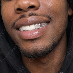 May include: A silver-toned dental grill featuring three circular settings, with the center setting containing a small, oval-shaped red gemstone. The grill is worn over the upper teeth, showcasing a metallic finish and intricate detailing on each circular frame.