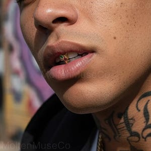 Pode incluir: Close-up da boca de uma pessoa com uma grelha de dentes de ouro. A grelha apresenta pequenas pedras preciosas coloridas. A pessoa tem uma tatuagem no pescoço e está vestindo uma blusa preta. A marca "MoltenMuseCo" é visível.