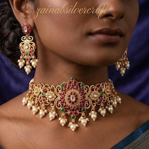 Op de afbeelding: Een goudkleurige ketting en bijpassende oorbellen set. De ketting heeft een centraal bloemmotief met rode en groene edelstenen, omringd door parels. De oorbellen hebben een vergelijkbaar ontwerp. De sieraden worden gepresenteerd tegen een donkerblauwe achtergrond.