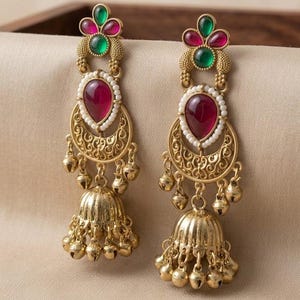 May include: A pair of gold-toned jhumka earrings featuring the text zainabsilvercraft. Each earring has a floral top with green and magenta stones, a central teardrop magenta stone surrounded by white beads, and a bell-shaped base with small hanging bells.