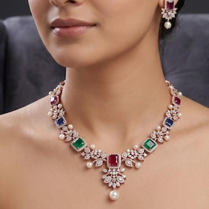 Op de afbeelding: Een ketting en bijpassende oorbellen van rosékleurig metaal met heldere pavé-stenen en afwisselend vierkant geslepen rode, blauwe en groene edelstenen. De sieraden bevatten kleine witte parels en een centrale hanger met een enkele parel.