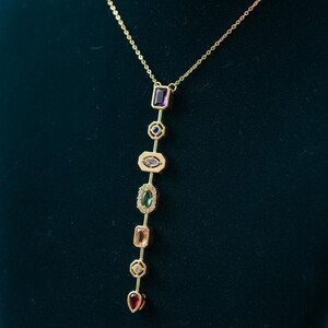 Può includere: Una collana color oro con una serie di pietre preziose colorate di varie forme. Le pietre preziose includono viola, blu, verde, pesca e rosso. La collana è esposta su uno sfondo verde scuro. Il testo "zainabsilvercrafts" è visibile.