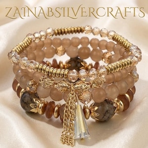Op de afbeelding: Stapel kralenarmbanden in bruin, beige en goudtinten. De armbanden hebben verschillende kraalgroottes, texturen en decoratieve elementen, waaronder een gouden kwast en de tekst "ZAINABSILVERCRAFTS".
