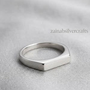 Puede incluir: Un anillo de metal en tono plateado pulido que presenta una superficie superior plana y rectangular. La banda tiene un acabado liso y continuo. El texto zainabsilvercrafts es visible en la parte superior derecha de la imagen, sobre el anillo.