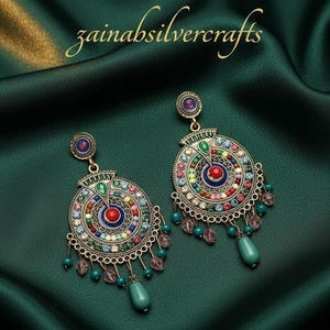 May include: A pair of ornate, colorful earrings with a circular design. The earrings feature a central red stone surrounded by concentric circles of various colored stones. Teal teardrop-shaped beads dangle from the bottom. The text "zainabsilvercrafts" is in the upper left.