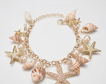 Op de afbeelding: Goudkleurige armband met schelp- en zeesterbedels, samen met imitatieparels. De armband bevat verschillende schelpvormen en -maten, met een karabijnsluiting. De tekst "zainabsilvercrafts" is zichtbaar bovenaan.