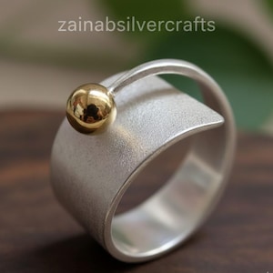 Könnte beinhalten: Ein silberner Ring mit einem goldfarbenen Kugelakzent. Der Ring hat ein breites Band mit strukturiertem Finish und geschwungenem Design. Die goldene Kugel sitzt auf einem gebogenen Silberband. Der Text "zainabsilvercrafts" ist sichtbar.