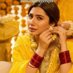 May include: A close-up of a woman in a yellow outfit, wearing red bangles and floral earrings. The background is blurred, featuring yellow flowers. The text "zainabsilvercrafts" is visible in the image.