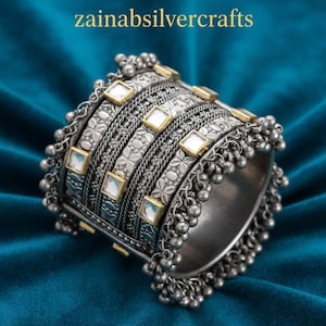 Peut inclure: Un bracelet manchette en argent avec des motifs floraux et géométriques complexes, orné de petits carrés blancs et de détails dorés. Le bracelet présente un design texturé et des perles d'argent pendantes. Le texte "zainabsilvercrafts" est visible.