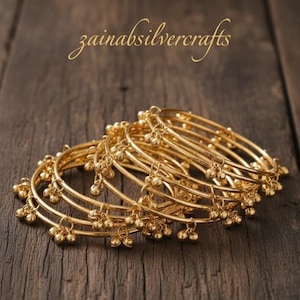 May include: A collection of gold-colored bangles with small, dangling bell-shaped charms. The bangles are stacked on a dark wooden surface. The text "zainabsilvercrafts" is in the upper left corner.