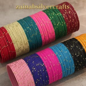 Pode incluir: Pilhas de pulseiras coloridas em vários tons, incluindo vermelho, rosa, verde e azul. As pulseiras são adornadas com pequenos pontos dourados. A imagem apresenta o texto "zainabsilvercrafts" no centro superior.