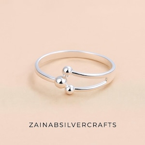 Puede incluir: Un anillo de plata con un diseño único. El anillo presenta una banda con tres pequeños acentos esféricos. El anillo se muestra sobre un fondo melocotón claro. El texto "ZAINABSILVERCRAFTS" está en la parte inferior de la imagen.