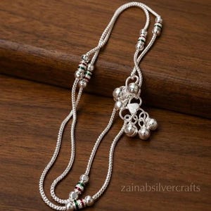 May include: A silver anklet featuring a delicate chain with small metallic beads and decorative accents in red and green. The anklet includes a cluster of dangling silver bells and a small heart-shaped charm. The text zainabsilvercrafts is visible.