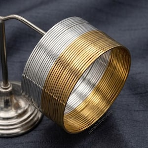 Può includere: Un set di braccialetti sottili e circolari disposti in pila, caratterizzati da sezioni alternate di metallo lucido color argento e oro. I braccialetti hanno un profilo liscio e arrotondato e sono esposti su un supporto in metallo dal design uniforme.