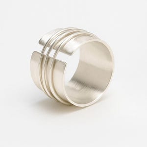 May include: A wide, silver ring with a contemporary design. The ring has a thick band with three parallel lines and cut-out sections. The text "ZAINABSILVERCRAFTS" is at the bottom. The ring is made of a shiny, metallic material.