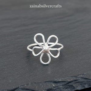 Puede incluir: Un anillo plateado con un diseño de flor de margarita. El anillo presenta una perla central y seis pétalos curvos. El texto "zainabsilvercrafts" está en la esquina superior izquierda.