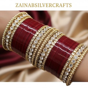 May include: An array of gold-coloured bangles, adorned with clear gemstones and a central band of deep red. The bangles are stacked on a wrist, highlighting the detailed design. The text "ZAINABSILVERCRAFTS" is visible at the top.