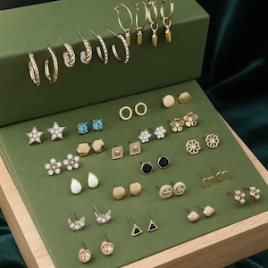 Könnte beinhalten: Eine Präsentation verschiedener goldfarbener Ohrringe, darunter Creolen, Ohrstecker und Hängeohrringe, angeordnet auf einer grünen Samtoberfläche. Die Ohrringe weisen verschiedene Designs auf, wie Sterne, Blumen und geometrische Formen. Der Markenname "zainabsilvercrafts" ist sichtbar.