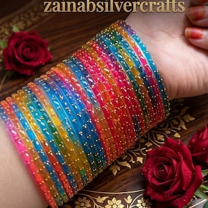May include: A vibrant stack of glass bangles in rainbow hues, including pink, orange, yellow, blue, and green, with gold-coloured detailing. The image also shows red roses and the text "zainabsilvercrafts".