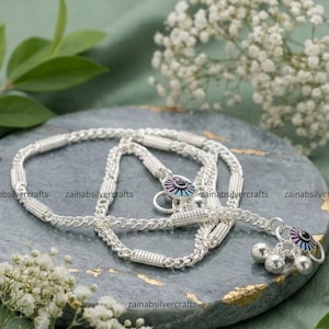 Può includere: Una cavigliera in argento con una catena a maglie cilindriche testurizzate alternate a segmenti intrecciati lisci. Il gioiello presenta piccoli campanellini pendenti e un ciondolo circolare con un centro scuro circondato da petali smaltati blu e viola.