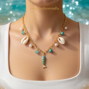 Può includere: Collana dorata con pendente a forma di pesce, conchiglie e dettagli turchesi e perle. La collana è indossata su un top bianco, con uno sfondo sfocato dell'oceano. Il testo "zainasilvercrafts" è visibile.