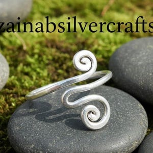 May include: A silver-toned ring with a spiral design. The ring features two swirling spirals connected by a band. The ring is displayed on a dark gray stone, with the text "zainabsilvercrafts" in black.
