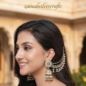 Puede incluir: Elegantes pendientes dorados con una piedra gris y perlas colgantes. Los pendientes presentan un diseño tradicional jhumka con detalles intrincados. El texto "zainabsilvercrafts" es visible en la parte superior.