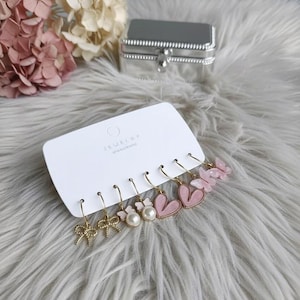 May include: A selection of six gold-coloured earrings on a white card. The earrings showcase bow, pearl, and heart designs in pink and gold. A silver jewellery box and pink flowers are in the background.