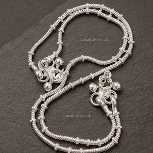May include: A pair of silver anklets featuring a delicate chain design with small, evenly spaced metallic beads along the length. Each anklet terminates in a cluster of small, round silver bells. The text zainabsilvercrafts is visible.