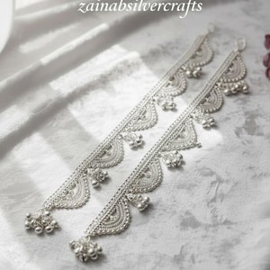 Puede incluir: Dos tobilleras plateadas con intrincados diseños festoneados y pequeñas campanillas colgantes. Las tobilleras se exhiben sobre una superficie blanca texturizada. El texto "zainabsilvercrafts" es visible.