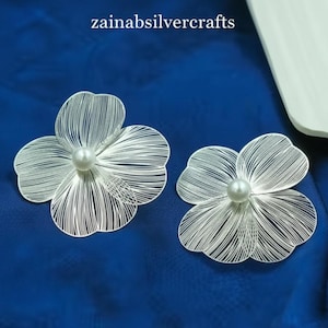 Könnte beinhalten: Zwei silberfarbene, blütenförmige Broschen mit Perlenmittelpunkt, vor tiefblauem Hintergrund. Die Broschen haben filigrane, lineare Designs, die Blütenblättern ähneln. Der Text "zainabsilvercrafts" ist oben sichtbar.