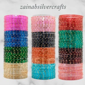 May include: Stacks of colorful bangles in shades of pink, blue, green, gold, black, orange, teal, red, and maroon. The bangles are transparent with gold accents. The text "zainasilvercrafts" is at the top of the image.