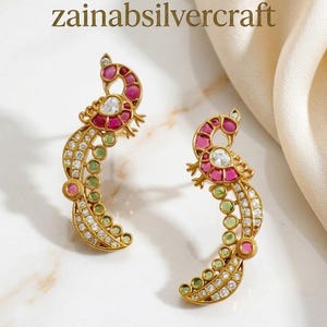 May include: A pair of gold peacock earrings. The earrings are adorned with red, green, and clear gemstones. The text "zainabsilvercraft" is visible. The earrings are displayed on a white surface.