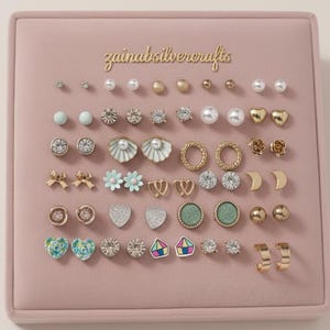 May include: A collection of stud earrings displayed on a pink velvet surface. The earrings feature various designs, including pearls, hearts, flowers, and geometric shapes. The text "zainabsilvercrafts" is in gold script at the top.