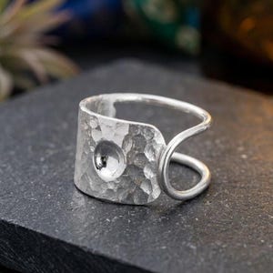 Könnte beinhalten: Ein silberfarbener Ring mit einem breiten, gehämmerten Metallband und einer konkaven, kreisförmigen Vertiefung in der Mitte. Das Band biegt sich zu einem offenen Design im Bypass-Stil, wobei ein glatter, abgerundeter Draht die Ringstruktur vervollständigt.
