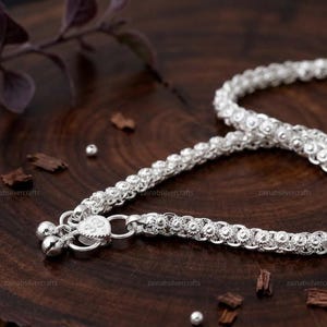 Könnte beinhalten: Ein silbernes Fußkettchen mit einem komplexen, geflochtenen Kettenmuster, einem herzförmigen Verschluss und zwei kleinen hängenden Glöckchen. Die metallische Oberfläche hat ein helles, reflektierendes Finish mit strukturierten Gliedern.