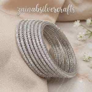 Puede incluir: Un conjunto de siete brazaletes de tono plateado, cada uno con una fila continua de pequeñas piedras claras y brillantes. El texto 'zainabsilvercrafts' es visible en la parte superior. Estos brazaletes circulares tienen un acabado metálico.