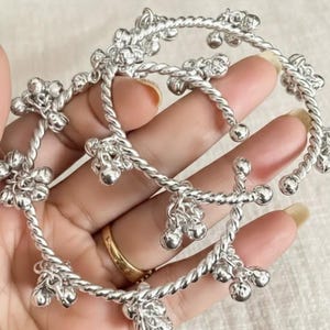 Op de afbeelding: Zilverkleurige armbanden met een gedraaid touwontwerp en kleine, bungelende belletjes. De armbanden worden in een hand gehouden, met de tekst "zainabs ilvercrafts" zichtbaar bovenaan. De sieraden hebben een heldere, reflecterende afwerking.