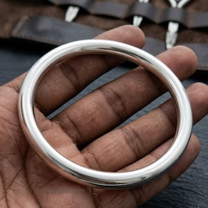 May include: A polished, circular silver bangle with a smooth, rounded surface and a reflective metallic finish. The bangle features a simple, continuous design without any additional embellishments, patterns, or textures, showcasing a clean, minimalist aesthetic.