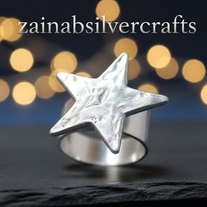 May include: A silver ring with a textured star design. The ring has a wide band and a large, five-pointed star. The text "zainabsilvercrafts" is visible in the background. The ring is set against a dark background with blurred lights.