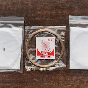May include: Three clear plastic bags with white contents and numbers 3, 0, and a package of bronze guitar strings. The center package has a red and white label with the brand name Ellora and the word BRONZE.